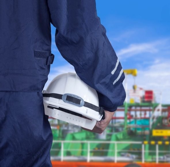 Engineer hand holding white safety helmet with blurred oil tanker in shipyard area